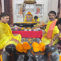 A traditional Hindu ritual scene inside a temple hall, with several men in bright yellow and orange robes seated around a decorated altar. A black Shivaling statue with orange marigold flowers is in the foreground, and a white carved deity niche with smaller offerings is in the background.